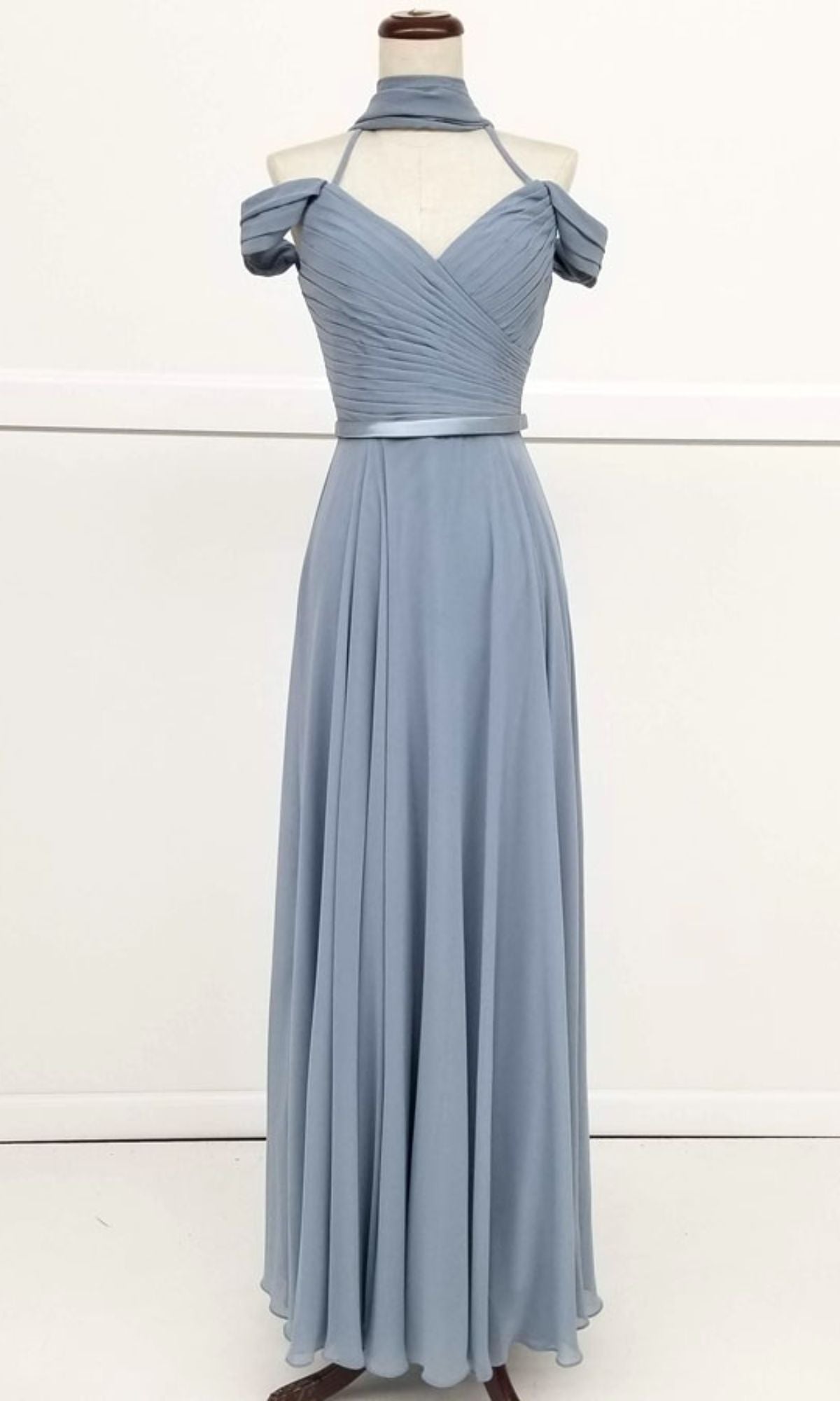 The Chicas C8997 Long A-Line Off-Shoulder Prom Dress is shown in light blue on a mannequin, featuring a ruched bodice, matching neck scarf, and flowing floor-length skirt.